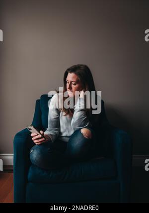 Attraktive Frau, die in einem bequemen Stuhl sitzt und ein Smartphone benutzt. Stockfoto