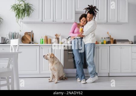 Fröhliches afroamerikanisches Paar, das sich in der Küche in der Nähe von labrador umarmt Stockfoto