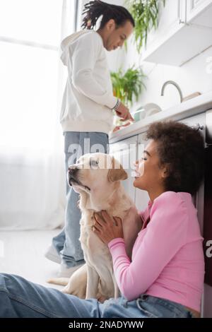 Seitenansicht einer lächelnden afroamerikanischen Frau, die labrador in der Nähe eines verschwommenen Freundes in der Küche umarmt Stockfoto