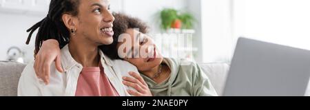 Lächelnde afroamerikanische Frau, die ihren Freund mit Dreadlocks umarmt, in der Nähe eines verschwommenen Laptops zu Hause, Banner Stockfoto