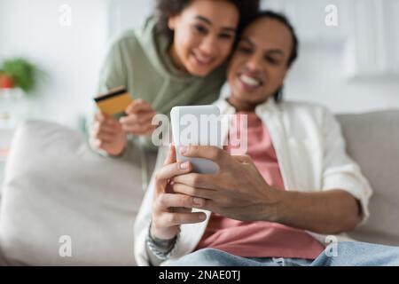 Verschwommenes afroamerikanisches Paar mit Smartphone und Kreditkarte beim Online-Shopping zu Hause Stockfoto