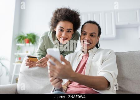 Lächelndes afroamerikanisches Paar, das beim Online-Shopping zu Hause ein Smartphone benutzte Stockfoto