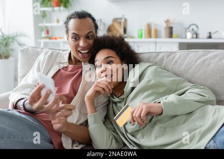 Aufgeregtes afroamerikanisches Paar, das Handy und Kreditkarte auf der Couch benutzte Stockfoto