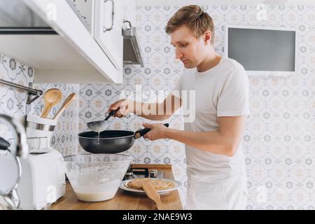 Ein junger Mann in Weiß, der Spaß hat, kocht Pfannkuchen Stockfoto