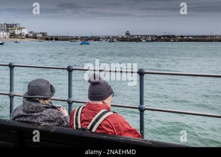 Wetter in Großbritannien. Besucher sitzen an einem regnerisch kalten, elenden Tag in der historischen Küstenstadt St. Ives in Cornwa auf einer Bank mit Blick auf den Smeatons Pier Stockfoto