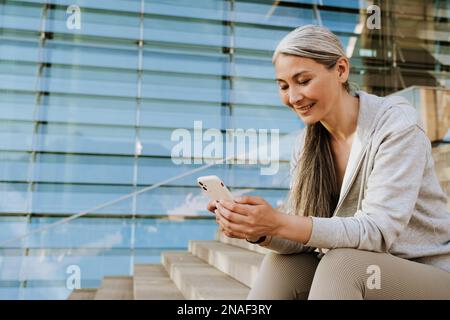 Asiatische reife Frau im Trainingsanzug, die ihr Handy benutzt und beim Workout im Freien auf Treppen sitzt Stockfoto