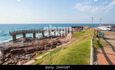 Angelpier Betonsteg an der felsigen Küste mit Blick auf das malerische blaue Meer am Sommertag mit Fischerfahrzeugen auf dem Parkplatz. Stockfoto