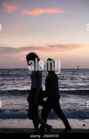Ein Paar mit Silhouette, das bei Sonnenuntergang am Strand spaziert; Melbourne, Victoria, Australien Stockfoto