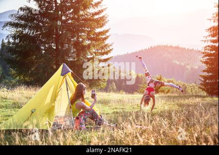 Zwei Frauen, die in den Bergen campen. Junge Frau auf Reisen, Wandern im Sommer. Konzentrieren Sie sich auf sportliche Frauen, die salto machen, andere sitzende Frauen, die Fotos machen. Konzept von Tourismus und Abenteuer. Stockfoto