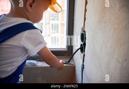 Nahaufnahme eines kleinen Bauarbeiters, der Drähte mit einer Zange in einer Wohnung schneidet, die gerade renoviert wird. Kind fixiert Elektrokabel in der Wand, während es an der Renovierung arbeitet. Stockfoto