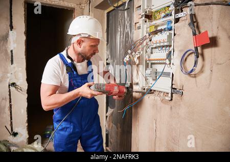 Männlicher Baumeister, der eine elektrische Schalttafel in der Wohnung installiert. Mann in Arbeitsanzug und Sicherheitshelm Bohrwand mit Elektrobohrmaschine, während er mit der Schalttafel an der Wand steht. Stockfoto