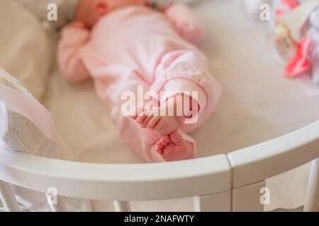 Kleine Babyfüße mit pinkfarbenem Strampelanzug. Kleine Finger des Neugeborenen. Das Kind hat sich die Beine gekreuzt und liegt im weißen Bett. Stockfoto