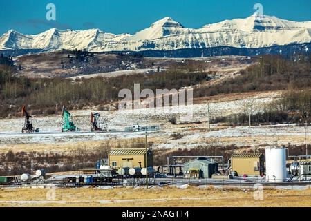 Kompressorstation mit Pumpjacks im Hintergrund, schneebedeckten Hang und Bergkette mit blauem Himmel, nördlich von Longview, Alberta Stockfoto