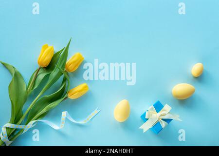 Osterfest-Hintergrund. Gelb bemalte Eier, Geschenkbox und Tulpen auf blauem Tisch. Draufsicht, flach liegend, Kopierbereich. Stockfoto