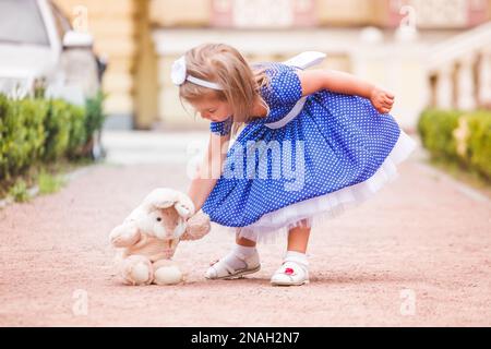 Das kleine Mädchen auf der Straße, das mit einem Spielzeug spielt. Baby legt Spielzeughase auf den Boden. Ein Mädchen in einem üppigen blauen Kleid, das in der Stadt Kiew (Ukraine) spaziert. Stockfoto