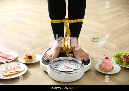 Essen, Alkohol nach Feiertagen und Frau mit Maßband, die drinnen auf Waage steht, Nahaufnahme. Übergewichtigkeitsproblem Stockfoto