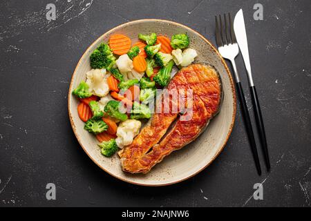 Gesundes Fischgericht: Gegrilltes Lachssteak mit Gemüsesalat auf Keramikplatte mit Gabel und Messer auf schwarzem Schieferstein von oben Stockfoto