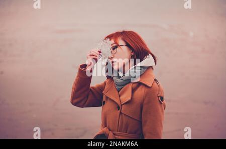 Pastellrosa Atmosphäre am Ufer. Eine schöne Asiatin steht in warmen Kleidern am Meer und hält durchsichtiges Eis in der Hand. Stockfoto