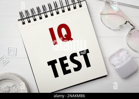 Notizbuch mit Text IQ Test, Brille und Ohrhörer auf weißem Holztisch, flach liegend Stockfoto