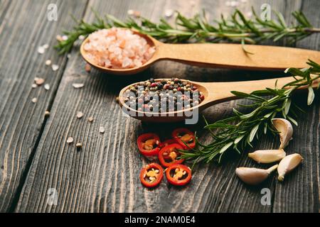 Nahaufnahme von gemischten Pfefferkörnern und Meersalz in Holzlöffeln, frischem Rosmarin und Knoblauch auf dunklem Holzhintergrund. Essen, kulinarisch, kochen Stockfoto