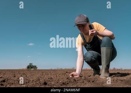 Landwirtin, die die Qualität des gepflügten Ackerbodens vor der Aussaat prüft, landwirtschaftlicher Traktor im Hintergrund, selektiver Fokus Stockfoto