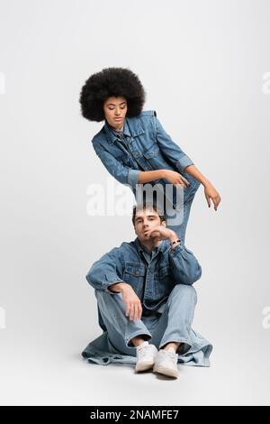 Die ganze Länge einer lockigen afroamerikanischen Frau steht hinter einem Mann in Jeansbekleidung und Turnschuhen auf grauem Stockbild Stockfoto