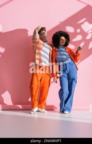 Trendige interrassistische Modelle in Sonnenbrille auf pinkfarbenem Hintergrund mit Schatten- und Stockbild Stockfoto