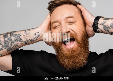 Porträt eines überfreulichen bärtigen Mannes, der den Kopf berührt und mit geschlossenen Augen lacht, isoliert auf einem grauen Stockbild Stockfoto