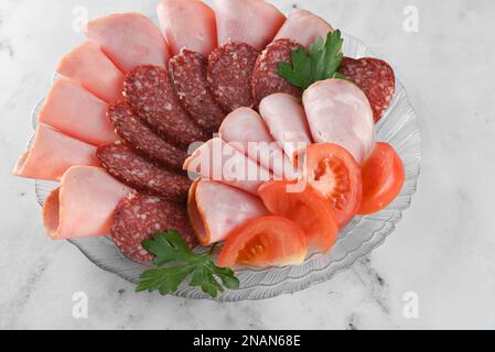 Teller mit einer Auswahl an gekochten, geräucherten Würstchen auf einem hellen Tisch. Wurstaufschnitt mit Kräutern und Gemüse Stockfoto