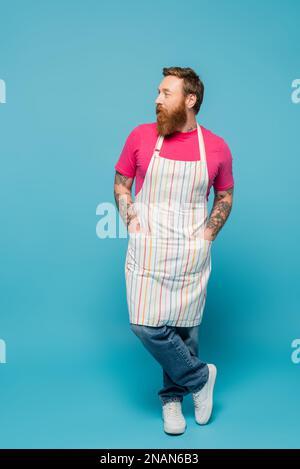 Die ganze Länge eines tätowierten Mannes, der mit Händen in Taschen mit gestreifter Schürze steht und auf blauen Hintergrund wegblickt, Stockbild Stockfoto