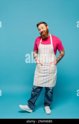 Fröhlicher bärtiger Mann, der mit Händen in Taschen mit gestreifter Schürze auf blauem Hintergrund posiert, Stockbild Stockfoto