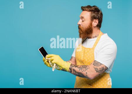 Bärtiger Mann in weißem T-Shirt und gelber Schürze, WischSmartphone mit leerem Bildschirm, isoliert auf blauem Stockbild Stockfoto