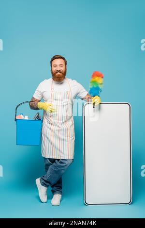 Fröhlicher bärtiger Mann in gestreifter Schürze mit Staubbürste und Eimer neben weißer Telefonvorlage auf blauem Hintergrund, Stockbild Stockfoto