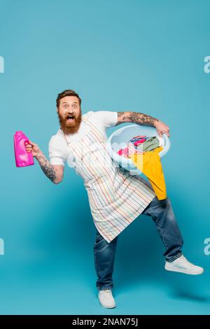 Die ganze Länge des aufgeregten bärtigen Mannes posiert mit Waschgel und Wäscheschale auf blauem Hintergrund, Stockbild Stockfoto