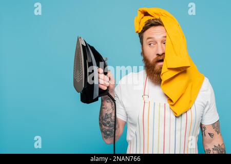 Bärtiger Mann mit gelbem Kleidungsstück auf dem Kopf, der auf elektrisches Eisen isoliert auf blauem Stock-Bild schaut Stockfoto