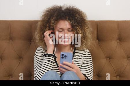 Glückliche junge Frau mit Locken, die Kopfhörer trägt und online Lieblingsmusik hört Stockfoto