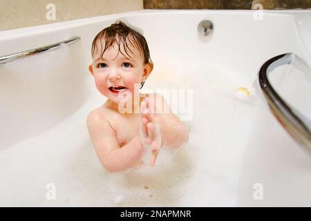 Ein glückliches Kind spielt mit Seifenblasen, während es in einer weißen Badewanne sitzt. Ein lächelnder kleiner Junge badet im Badezimmer. Ein Kind im Alter von einem Jahr Stockfoto