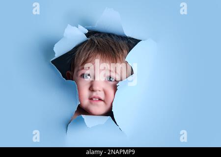 Trauriges Baby in einem Loch auf papierblauem Hintergrund. Zerrissener Studiohintergrund des Kindes, Kopierraum. Ein Kind im Alter von einem Jahr, sechs Monaten Stockfoto