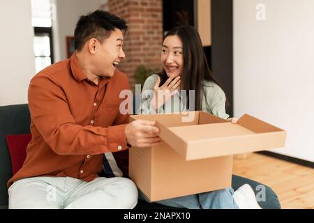 Überraschtes koreanisches Paar, das Kartonpakete ausgepackt hat, nachdem es erfolgreich eingekauft hatte, und zu Hause auf dem Sofa im Wohnzimmer saß Stockfoto