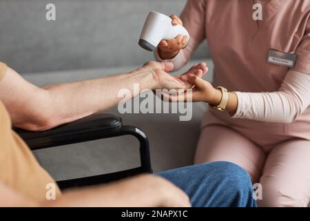 Nahaufnahme der Pflegekraft im gleichförmigen Haltearm des Seniorenmannes und Temperaturmessung mit spezieller medizinischer Ausrüstung Stockfoto