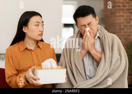 Ein kranker asiatischer Mann, eingewickelt in eine Decke, sitzt auf dem Sofa und niest die Nase, eine junge Frau behandelt ihren kranken Mann mittleren Alters Stockfoto