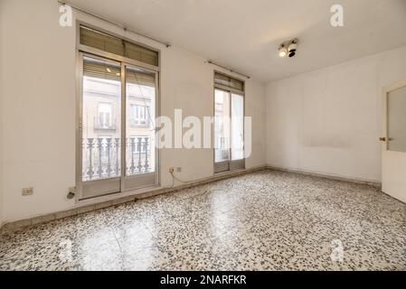 Leeres Wohnzimmer mit weißem Terrazzo-Boden mit schwarzen Punkten und Schiebetüren aus Aluminium und Saldia-Glas zu zwei kleinen Balkonen mit schwarzem Metallgeländer Stockfoto