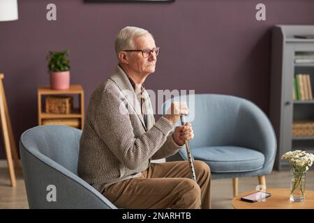 Senior-Mann mit Stock, sitzt auf einem Sessel mit nachdenklichem Anblick und verbringt Zeit zu Hause Stockfoto