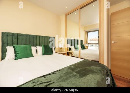 Schlafzimmer mit einem Doppelbett mit grünen Kissen und einem gepolsterten Kopfteil in der gleichen Farbe neben einem Kleiderschrank mit Spiegelschiebetüren Stockfoto