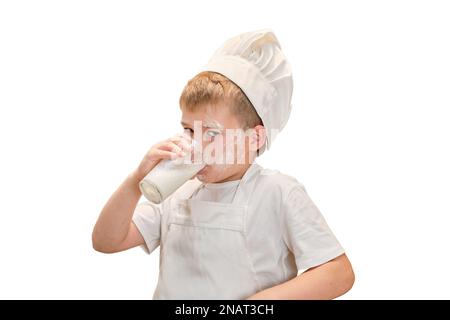 Ein Junge in der Kleidung des Küchenchefs trinkt Milch aus einem Glas, während er in der Küche einen Apfelkuchen kocht Stockfoto