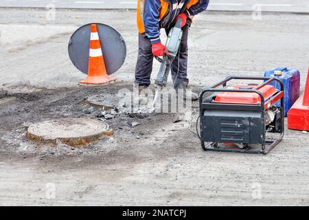 Ein Straßenarbeiter zerstört alten Asphalt in der Nähe eines Kanalschachts mit einem elektrischen Presslufthammer. Stockfoto