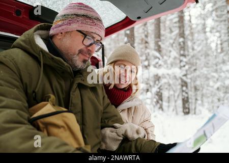 Seniorenpaar in warmer Winterbekleidung, schaute sich eine Karte an, die ein Mann in Papierform hielt, während beide auf dem Kofferraum saßen, während eine Gruppe von Bäumen mit Schnee bedeckt war Stockfoto