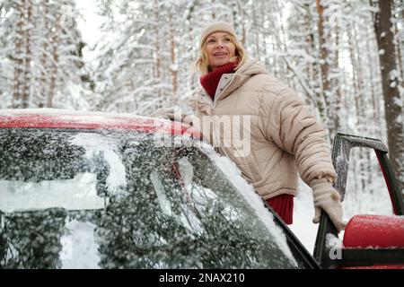 Glückliche, reife blonde Frau in warmer Winterjacke, gestrickter Beanie und Wollhandschuhe, die während der Reise an der offenen Tür des Autos stand Stockfoto