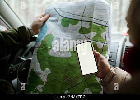 Nahaufnahme der Hand einer erwachsenen Frau, die ihr Smartphone über die Karte ihres Mannes hielt, während sie im Winter mit dem Auto unterwegs war Stockfoto