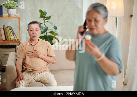 Älterer Mann, der an plötzlichem Herzschmerz leidet, als seine Frau einen Arzt anruft, um zu fragen, welches Medikament er einnehmen soll Stockfoto